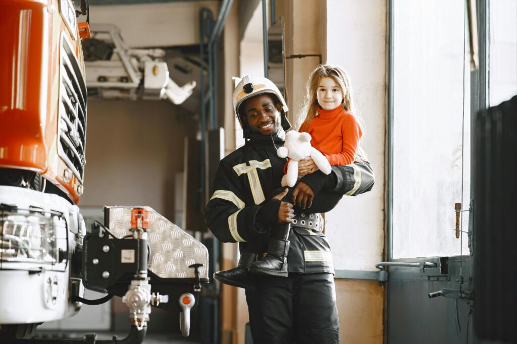 Firefighter holding child with teddy bear, near fire truck.